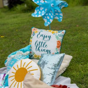 Colorful pinwheel with blue and white polka-dotted blades, set against a grassy outdoor background with decorative pillows and a sign that reads "Enjoy the little things.".