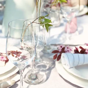 An elegant outdoor table setting with clear wine glasses, white plates, pink floral napkins, and a white tablecloth, with greenery in the background.