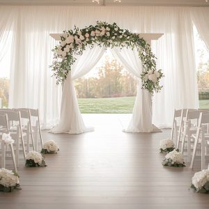 Elegant outdoor wedding ceremony setup with a floral arch, white drapes, and rows of white chairs on a wooden floor, overlooking a scenic landscape.