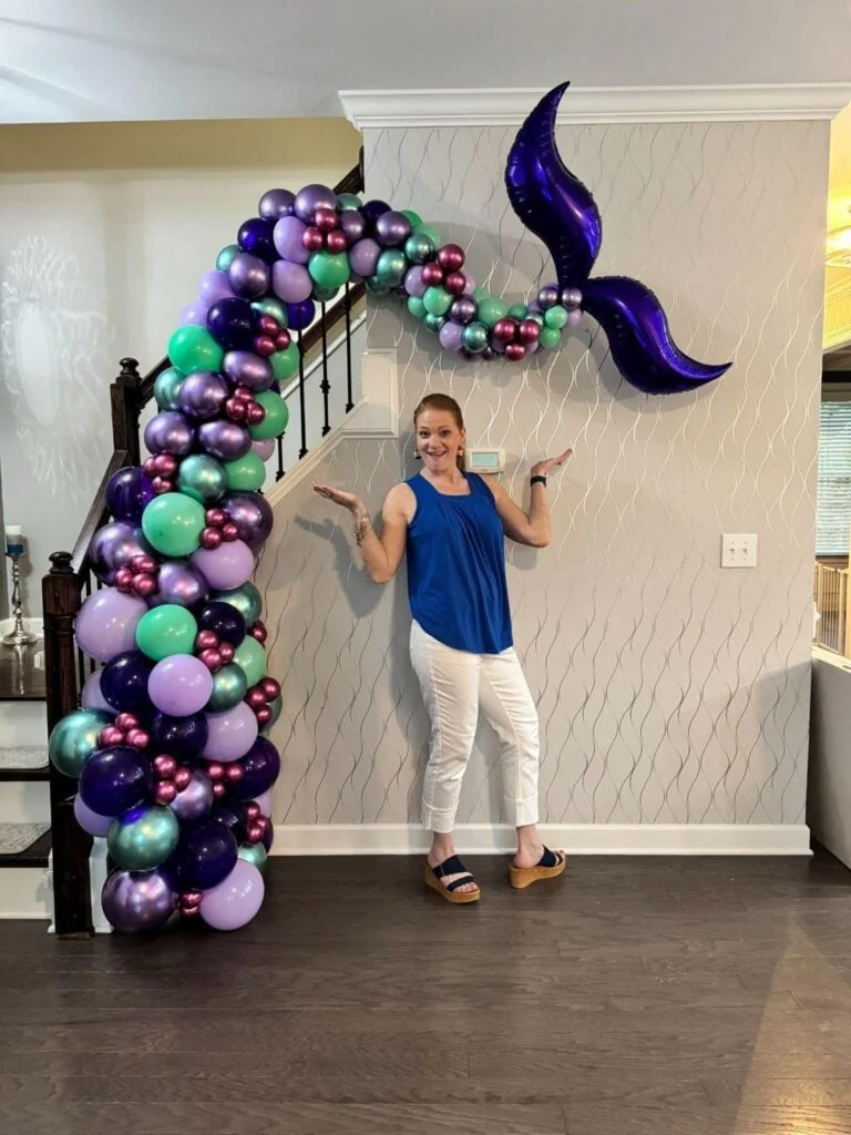 Woman standing next to a balloon arch with purple, teal, and pink balloons and a large purple balloon shaped like a crescent moon, indoors against a patterned wall.