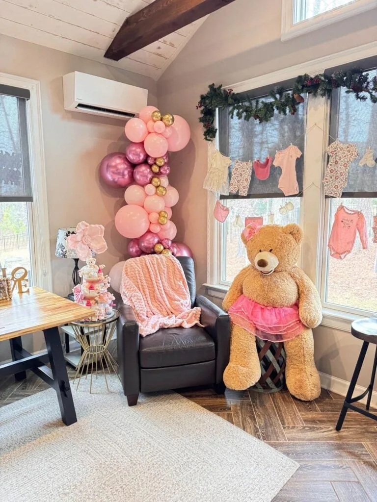 Cozy corner with a large teddy bear dressed in a pink tutu, pink and gold balloon decoration, and a garland of children's clothes hanging by the window.