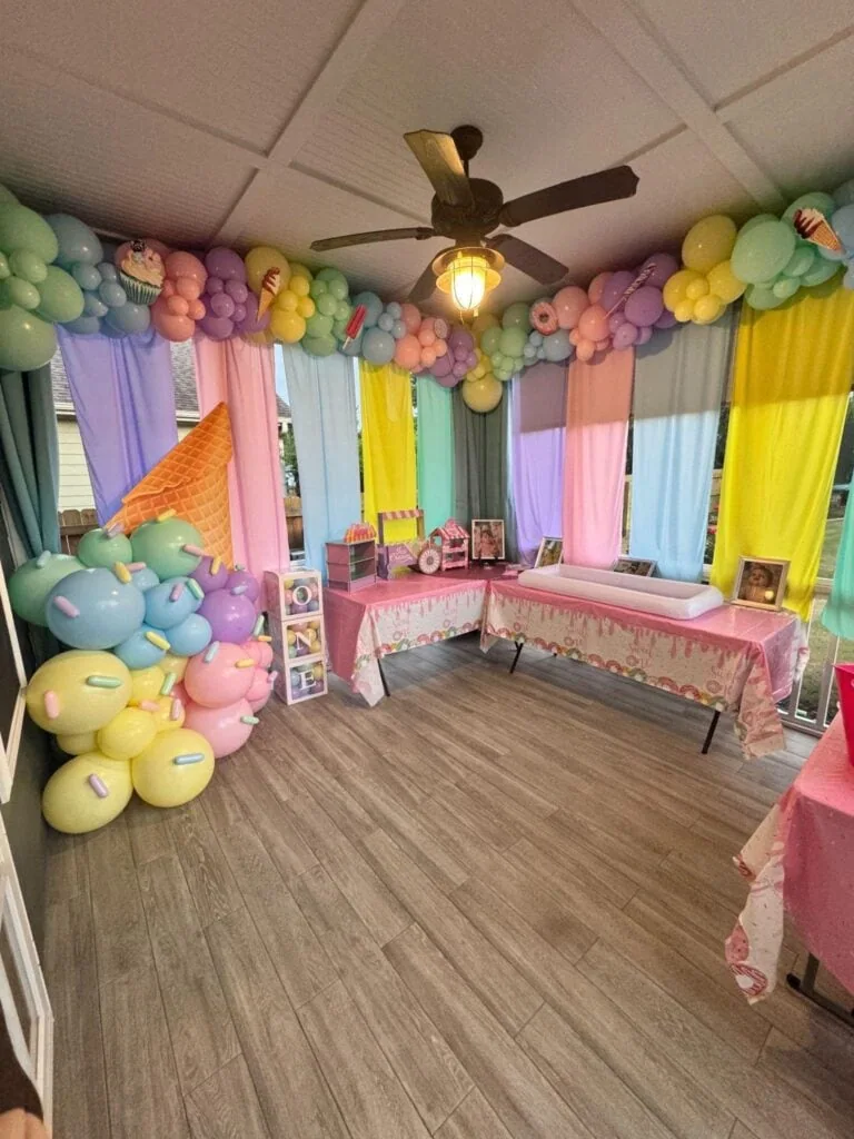 A colorful birthday party room decorated with pastel balloons, curtains, and themed table settings, featuring a ceiling fan and wooden flooring.