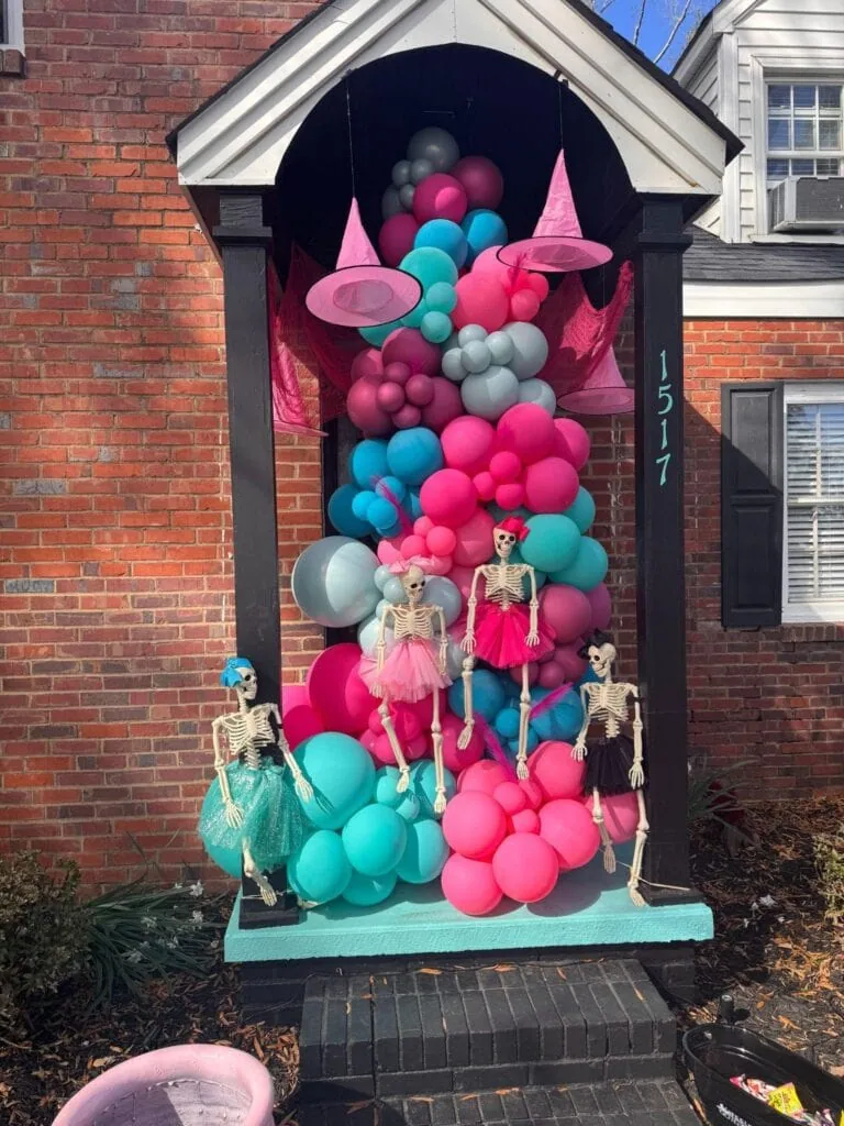 Pink, blue, and gray balloons arranged in a whimsical display with skeleton mannequins dressed in colorful outfits, set against a brick house entrance with the number 1517.