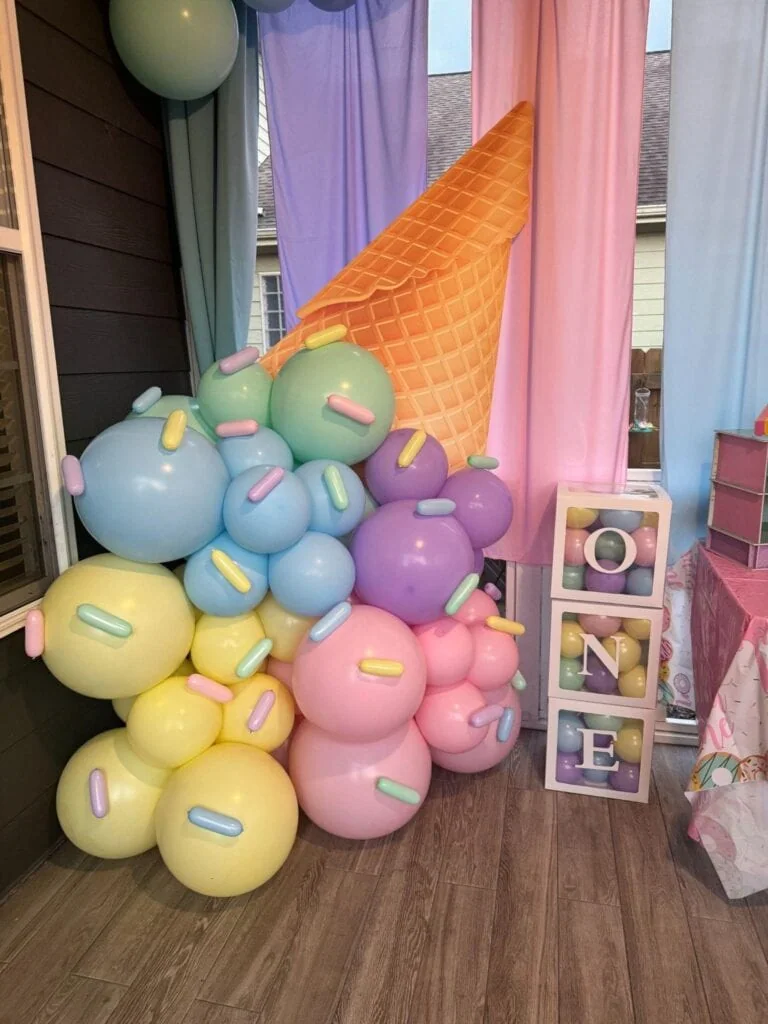 Colorful balloon arrangement resembling an ice cream cone with pastel balloons in pink, purple, yellow, and blue, and a large cone prop, set against pastel curtains and a small decorative cube with th.
